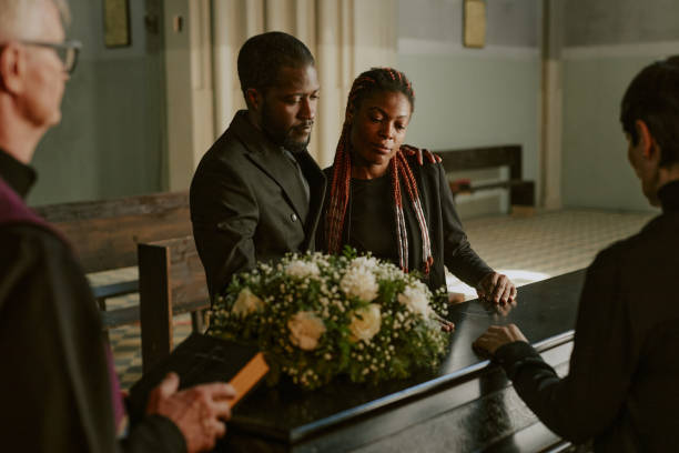 African American husband and wife looking at black coffin and bemoaning their loss together with senior pastor and their family friend